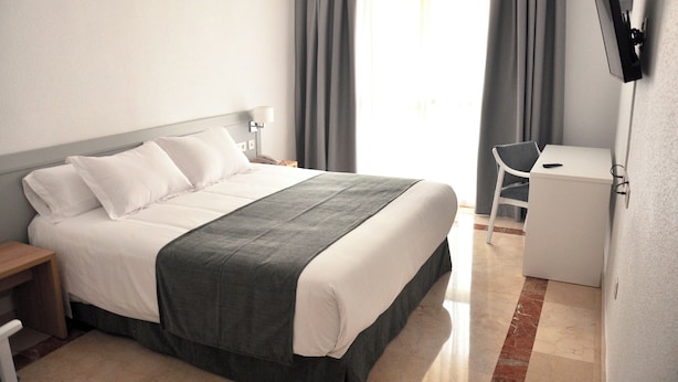 Una habitación de hotel con cama, escritorio, silla, televisor montado en la pared y ventana con cortinas.