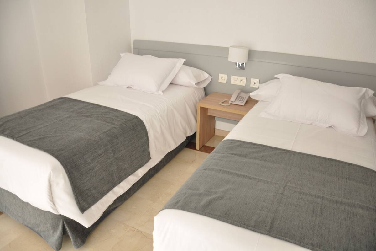 Quarto de hotel com duas camas, cada uma com um cobertor cinzento e almofadas brancas. Há uma mesa de cabeceira de madeira com uma luminária branca e um telefone.
