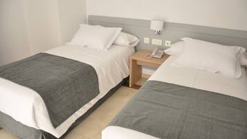 Una habitación de hotel con dos camas, cada una con una manta gris y almohadas blancas. Hay una mesita de noche de madera con una lámpara blanca y un teléfono.