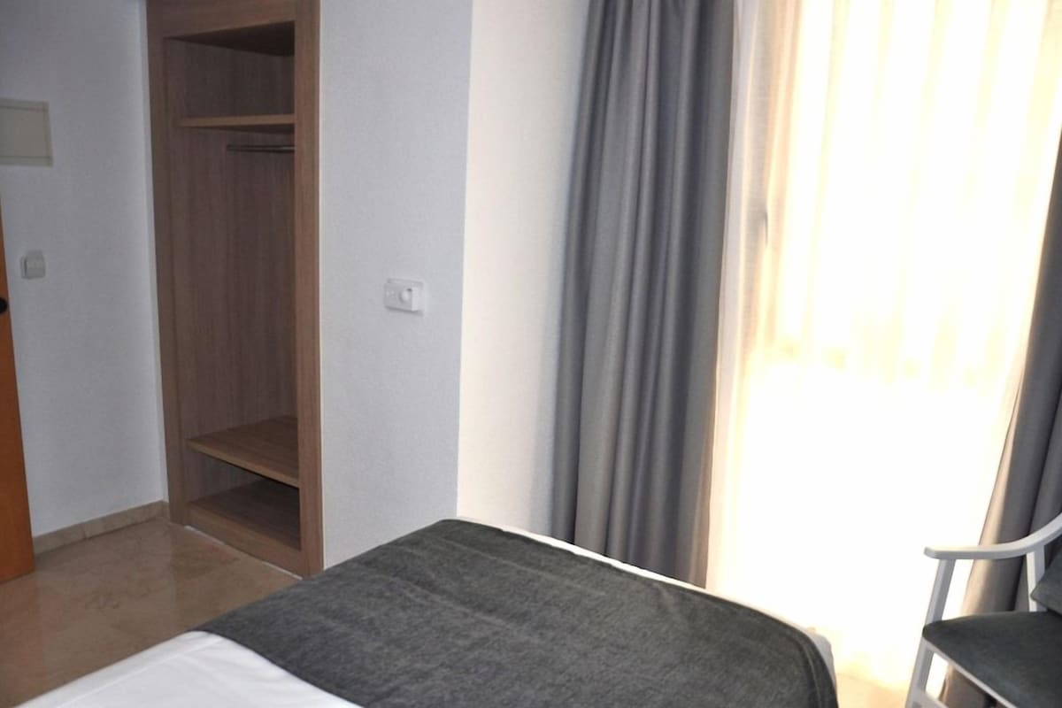A hotel room with a bed, a chair, a wardrobe, and a window with curtains.