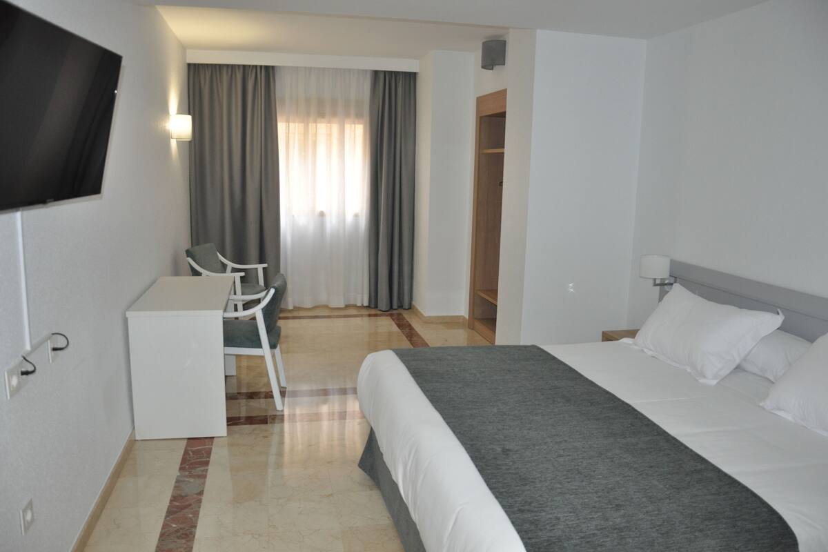 Quarto de hotel com cama, escrivaninha, cadeira, televisão e guarda-roupa.
