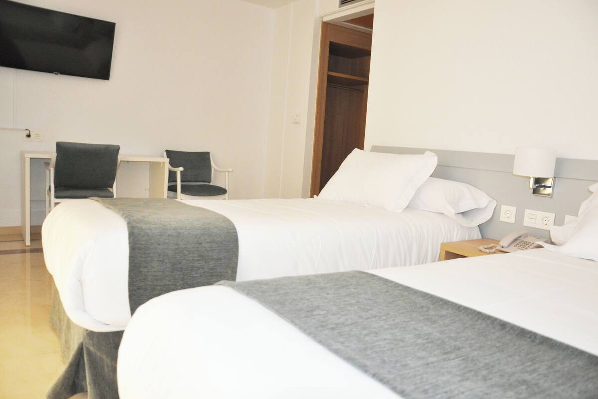 Quarto de hotel com duas camas, televisão presa à parede, escrivaninha com cadeira e guarda-roupa.