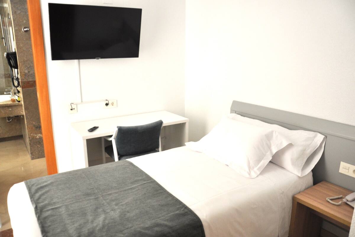 Quarto de hotel com uma cama, uma escrivaninha, uma cadeira, uma televisão e um telefone.