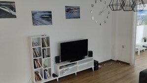 A modern living room with a flat-screen TV on a white entertainment unit, a bookshelf filled with books, and a wall clock. - Salimana charming apartman (Novi Sad)