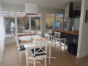 Un coin repas avec des chaises blanches et une table en bois, un espace cuisine avec un poêle noir, et de grandes fenêtres offrant une vue sur l’extérieur.