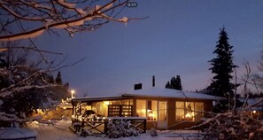 Une maison recouverte de neige, dotée d’une terrasse en bois et dont l’intérieur est éclairé.