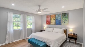 A bedroom with a bed, a ceiling fan, a bench, two bedside tables, and a large window with curtains. - Family Game House Near Kings Island (Mason)