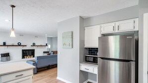 A modern kitchen with white cabinets, a stainless steel refrigerator, a microwave, and a built-in oven. There is a blue sofa in the adjacent living area, and a fireplace with candles. - Family Game House Near Kings Island (Mason)