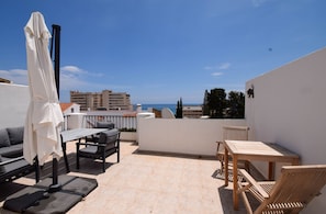 A rooftop terrace with outdoor furniture, a table, and a view of the sea and buildings.