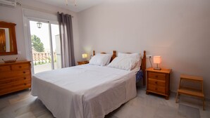 Een slaapkamer met een groot bed, een houten dressoir, een nachtkastje met een lamp, een kleine tafel en een plafondventilator.