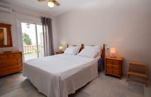 A bedroom with a large bed, a wooden dresser, a nightstand with a lamp, a small table, and a ceiling fan.