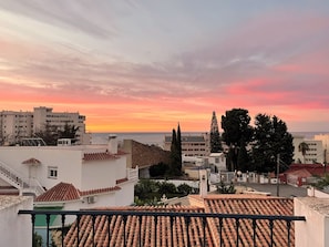 Un paysage urbain côtier au coucher du soleil, avec des bâtiments aux toits de tuiles rouges, un ciel dégagé et une vue sur la mer.
