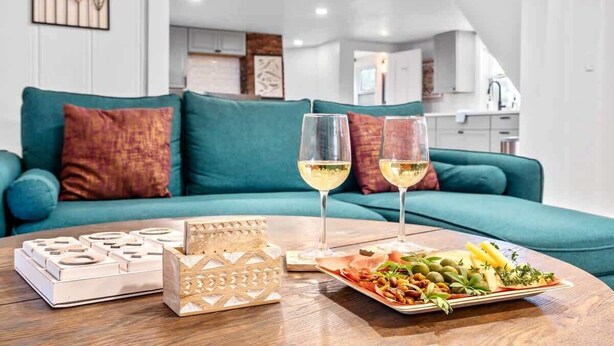 A modern living room with a teal sofa, two glasses of white wine, a tray of food, and a decorative candle on a wooden table. - Peaceful 1BR | Reading Nook | Near Portland (Westbrook)
