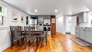 A modern kitchen with grey cabinets, a brick accent wall, and a dining area with wooden chairs. - Peaceful 1BR | Reading Nook | Near Portland (Westbrook)