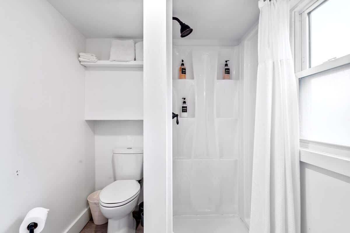 Un baño pequeño y blanco con inodoro, un estante con toallas dobladas y una zona de ducha con cortina.