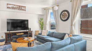 A modern living room with a sectional sofa, a flat-screen TV, a wooden coffee table, and a cityscape painting on the wall. - Downtown Penthouse Loft with Roof Patio and free parking (Louisville)