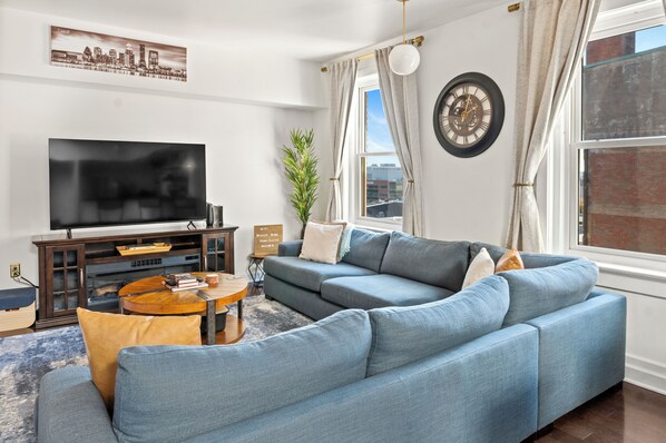 A modern living room with a sectional sofa, a flat-screen TV, a wooden coffee table, and a cityscape painting on the wall.