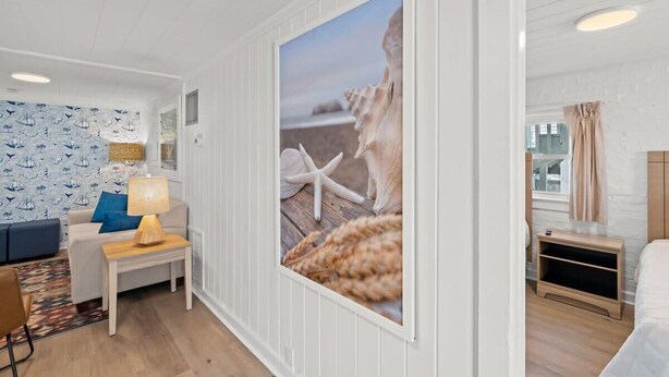 A room with a white wall, a framed picture of seashells, a blue patterned wallpaper, a wooden side table with a lamp, a blue sofa, and a bed with a wooden headboard. - Pet-Friendly, Steps to the Beach Seahorse Ranch #1 (North Myrtle Beach)