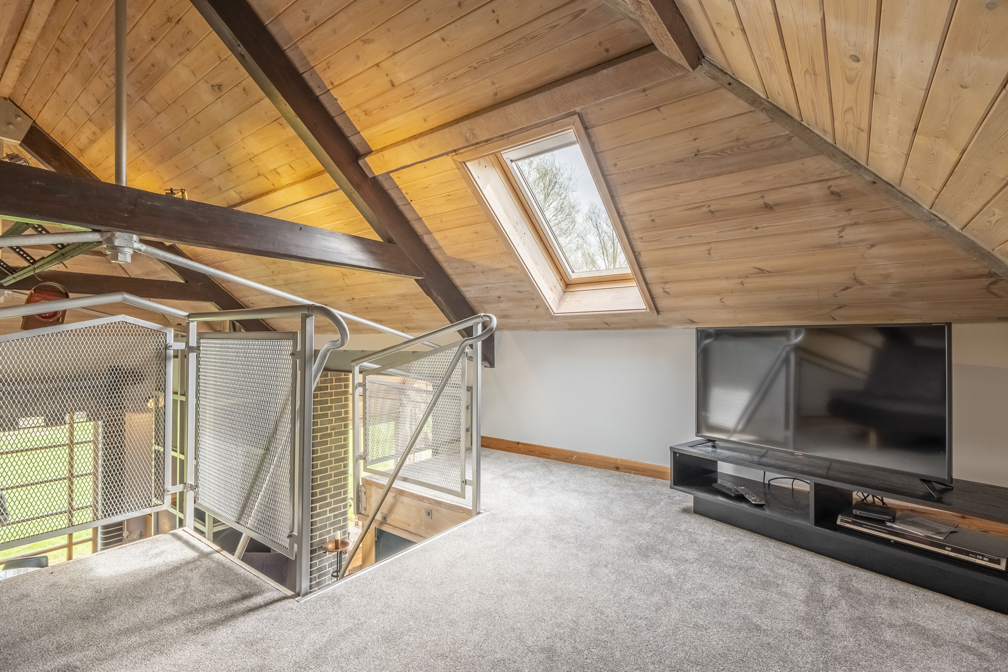 A high-ceilinged attic room with a skylight, a staircase with metal railings, and a TV stand with a flat-screen TV.