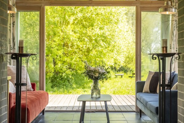 A room with a bed, a sofa, a small table with a vase of flowers, and a view of a green garden through large windows.