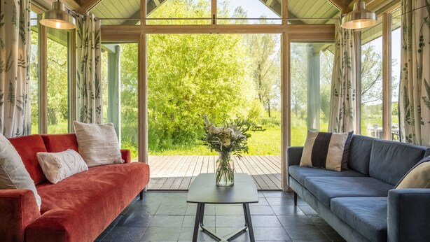 Ein gemütliches Zimmer mit einem rot-blauen Sofa, einem Glastisch und Blick auf einen üppigen Garten durch große Fenster.