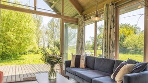 A modern living room with a grey sofa, a small table with a vase of flowers, and large windows offering a view of a green garden. - Ultimate Constable Country retreat -The Pump House (Colchester)
