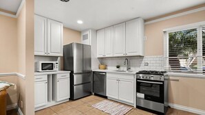 A kitchen with white cabinets, a stainless steel refrigerator, a microwave, a stove, and a window with blinds.