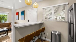 A kitchen with a white countertop, two bar stools, a stainless steel refrigerator, and a window with blinds.