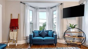 Ein modernes Wohnzimmer mit einem blauen Sofa, einem großen Fenster mit weißen Vorhängen, einem Flachbildfernseher und einem Holzbücherregal.