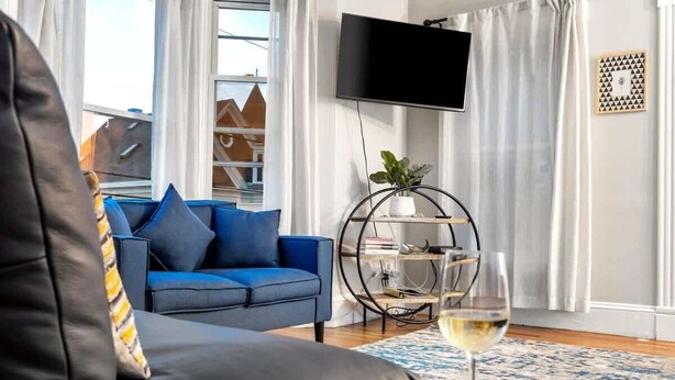 Ein modernes Wohnzimmer mit einem blauen Sofa, einem schwarzen Ledersessel und einem Glas Weißwein auf einem Holztisch.