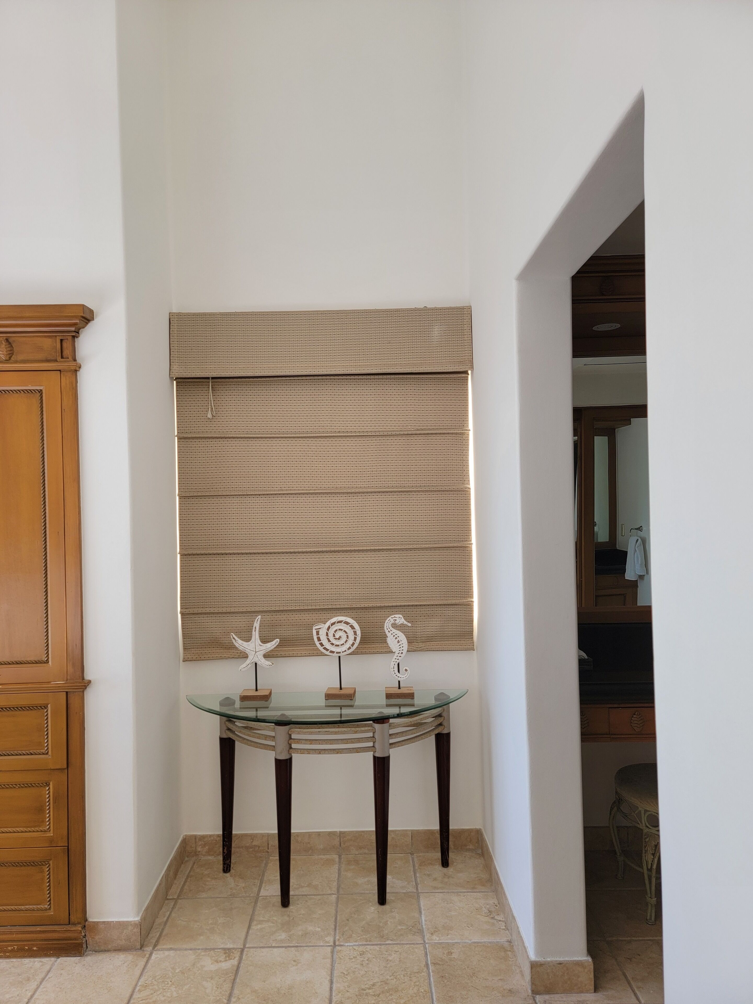 Un couloir avec une table en verre sur laquelle sont exposés trois objets décoratifs, un store beige à motifs recouvrant une fenêtre et un meuble en bois sur la gauche.