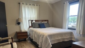 A bedroom with a bed, bedside table, lamp, chair, and a window with curtains. - Deer Ridge Duplex - Beautiful Lake View with Shared Dock! (Sunrise Beach)