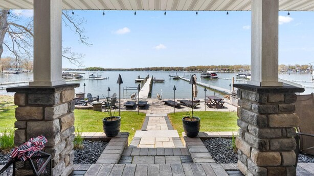 A covered outdoor area with stone pillars, a paved walkway, lounge chairs, and potted plants.