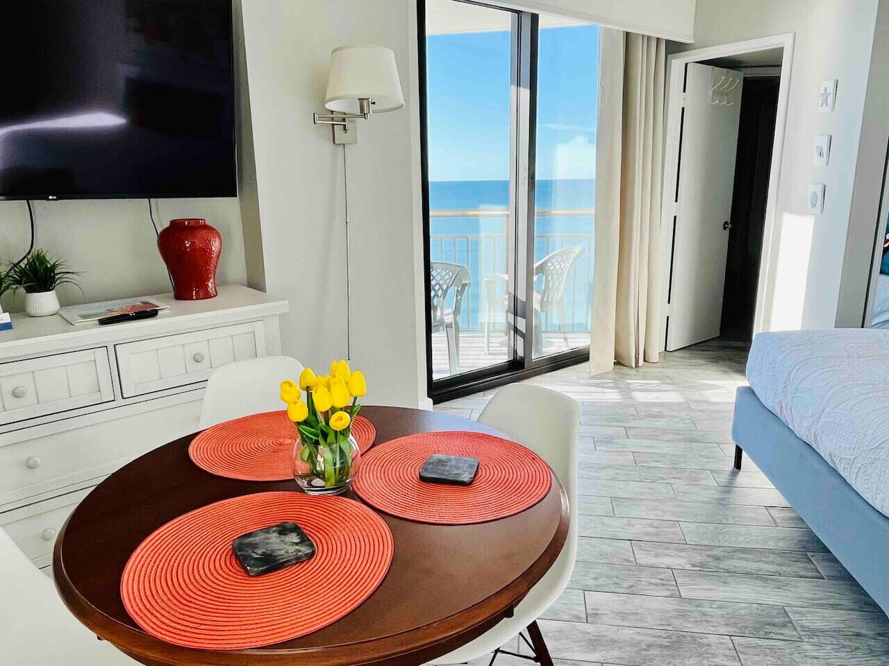 Une chambre d’hôtel moderne dotée d’un balcon, d’un coin repas avec une table et des chaises, d’un lit et d’un téléviseur fixé au mur.
