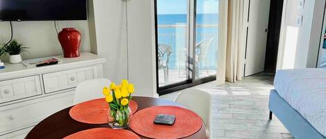Une chambre d’hôtel moderne dotée d’un balcon, d’un coin repas avec une table et des chaises, d’un lit et d’un téléviseur fixé au mur.