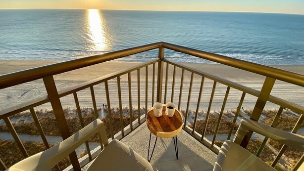Un balcón con una mesa de madera y dos tazas, con vista a una playa donde el sol se pone sobre el océano.