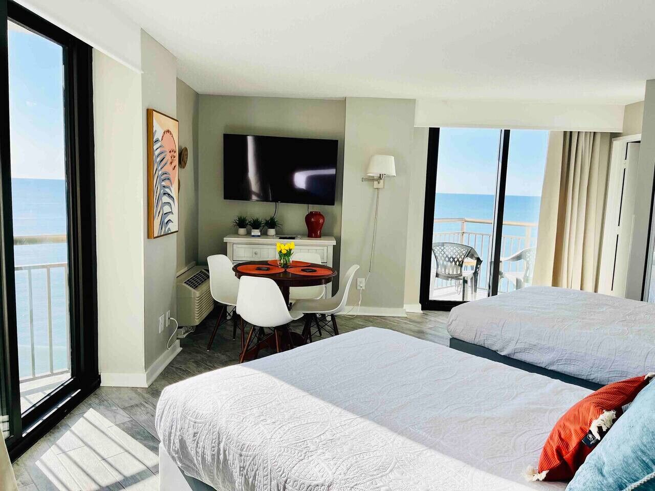 A hotel room with a balcony, a TV, a desk with chairs, and two beds.