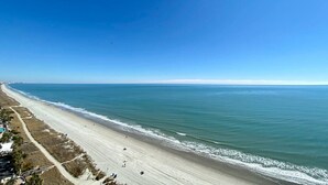 Una playa costera con cielo despejado, una orilla arenosa y algunas personas caminando.