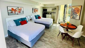 Habitación de hotel con dos camas, una mesa de comedor con sillas y una kitchenette al fondo.