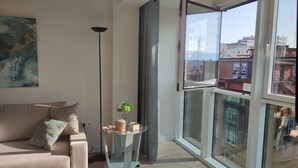 Ein Wohnzimmer mit einer Couch, einem kleinen Tisch mit einer Pflanze, einer Stehlampe und einem großen Fenster mit Blick auf Gebäude.