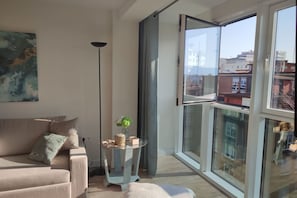 A living room with a sofa, a small table with a plant, a floor lamp, and a large window with a view of buildings. - 178A Mar de Gijón: Apartment with Views by R2R (Gijón)