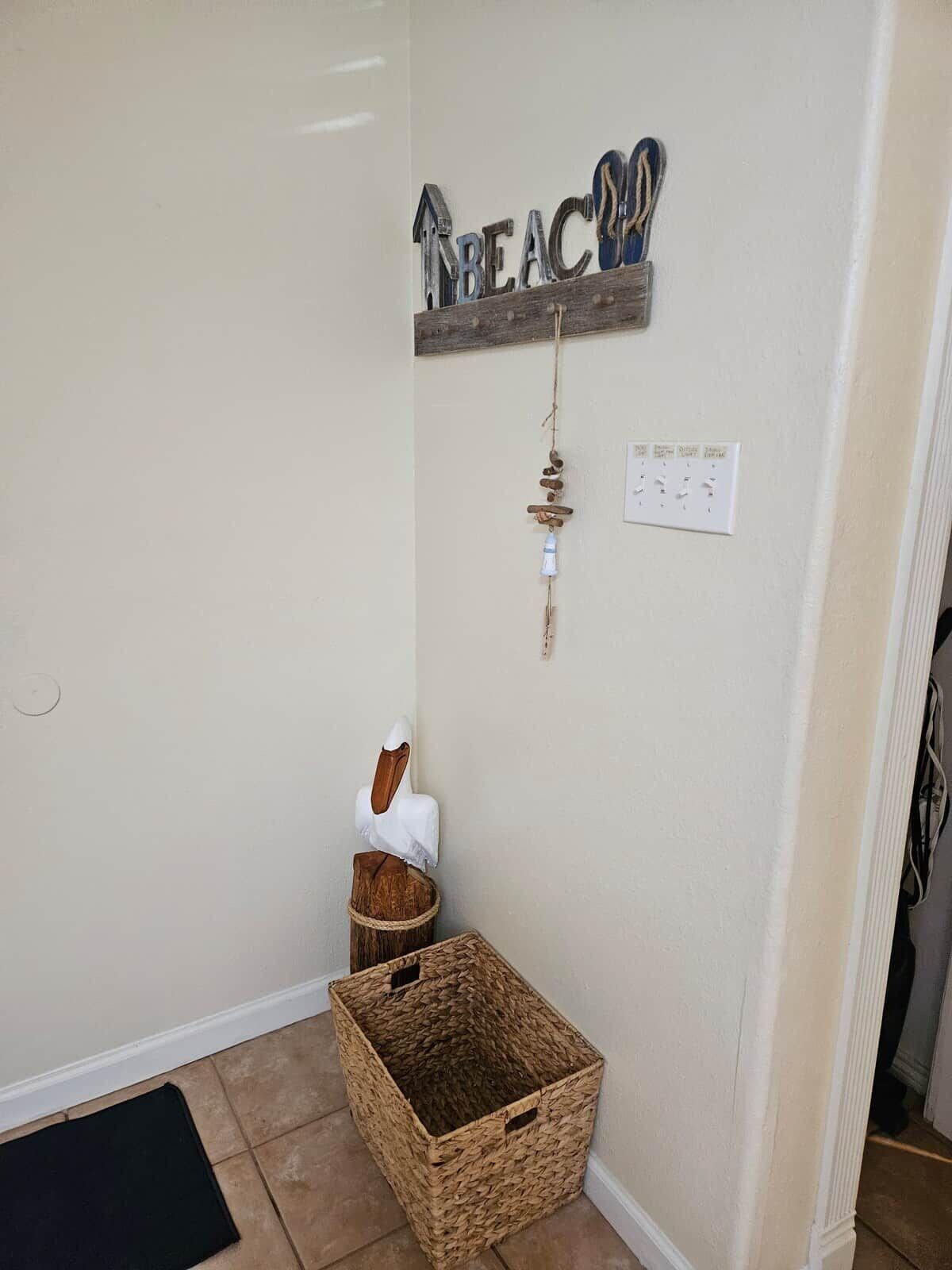 Una esquina de una habitación con una cesta tejida, una lámpara fijada a la pared y un letrero decorativo que dice 'BEAC'.