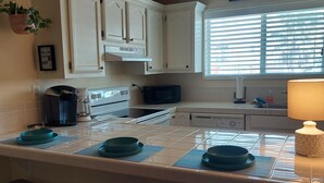 Cocina con armarios blancos, microondas, estufa y ventana con persianas. Hay dos taburetes y un letrero en la pared que dice 'Good Food Good Friends Good Times'.