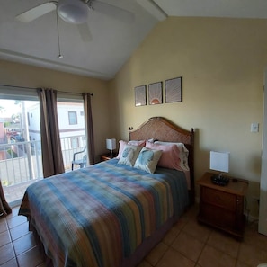 A bedroom with a bed, a nightstand, a ceiling fan, and a view of the outside.