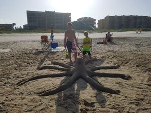 A sandy beach with a large octopus sand sculpture, buildings in the background, and a few people.