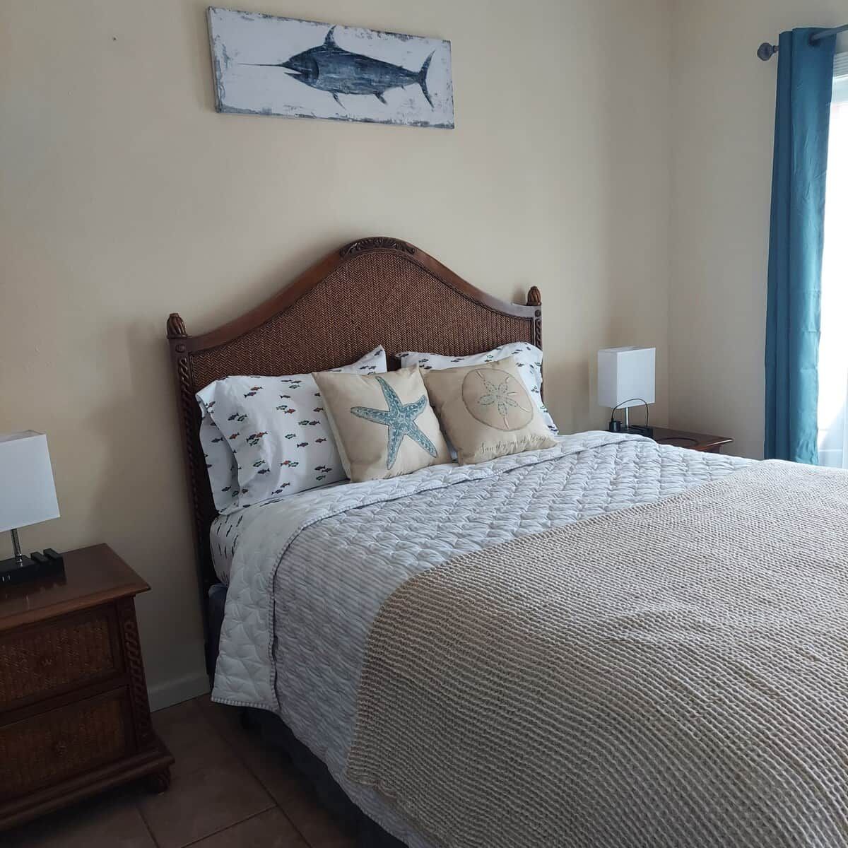 Una cama bien hecha con una colcha estampada, dos almohadas con diseños de estrellas de mar y conchas marinas, un cabecero de madera y un cuadro colgado en la pared con un pez.