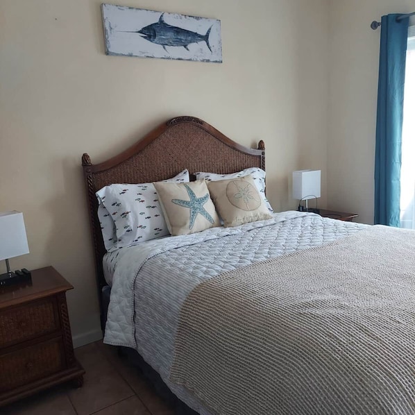 A neatly made bed with a patterned bedspread, two pillows with starfish and seashell designs, a wooden headboard, and a wall-mounted picture of a fish.