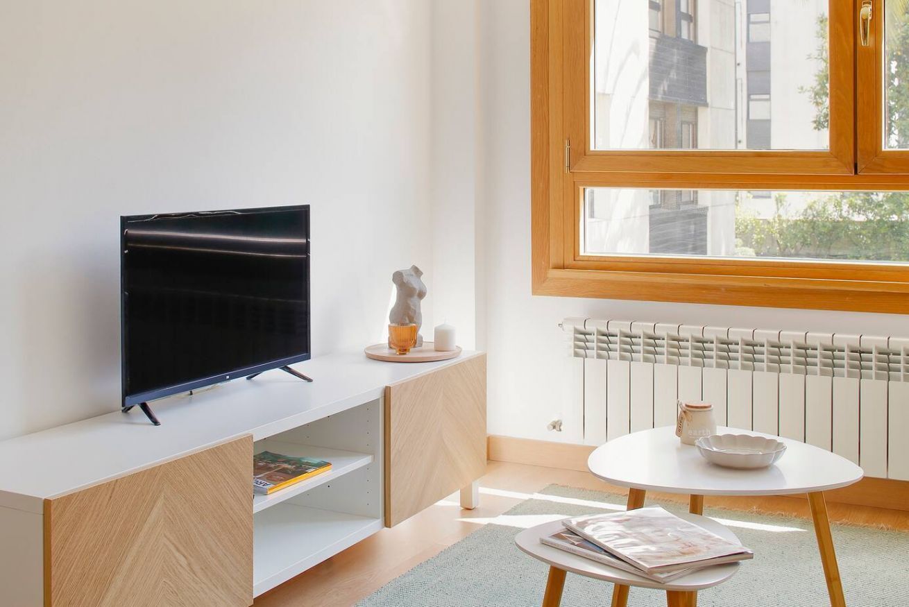 Un soggiorno moderno con una TV a schermo piatto su un mobile bianco, un tavolino con una ciotola e una tazza, e una finestra che offre vista su edifici.