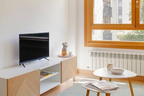 A modern living room with a flat-screen TV on a white cabinet, a small table with a bowl and a cup, and a window with a view of buildings.