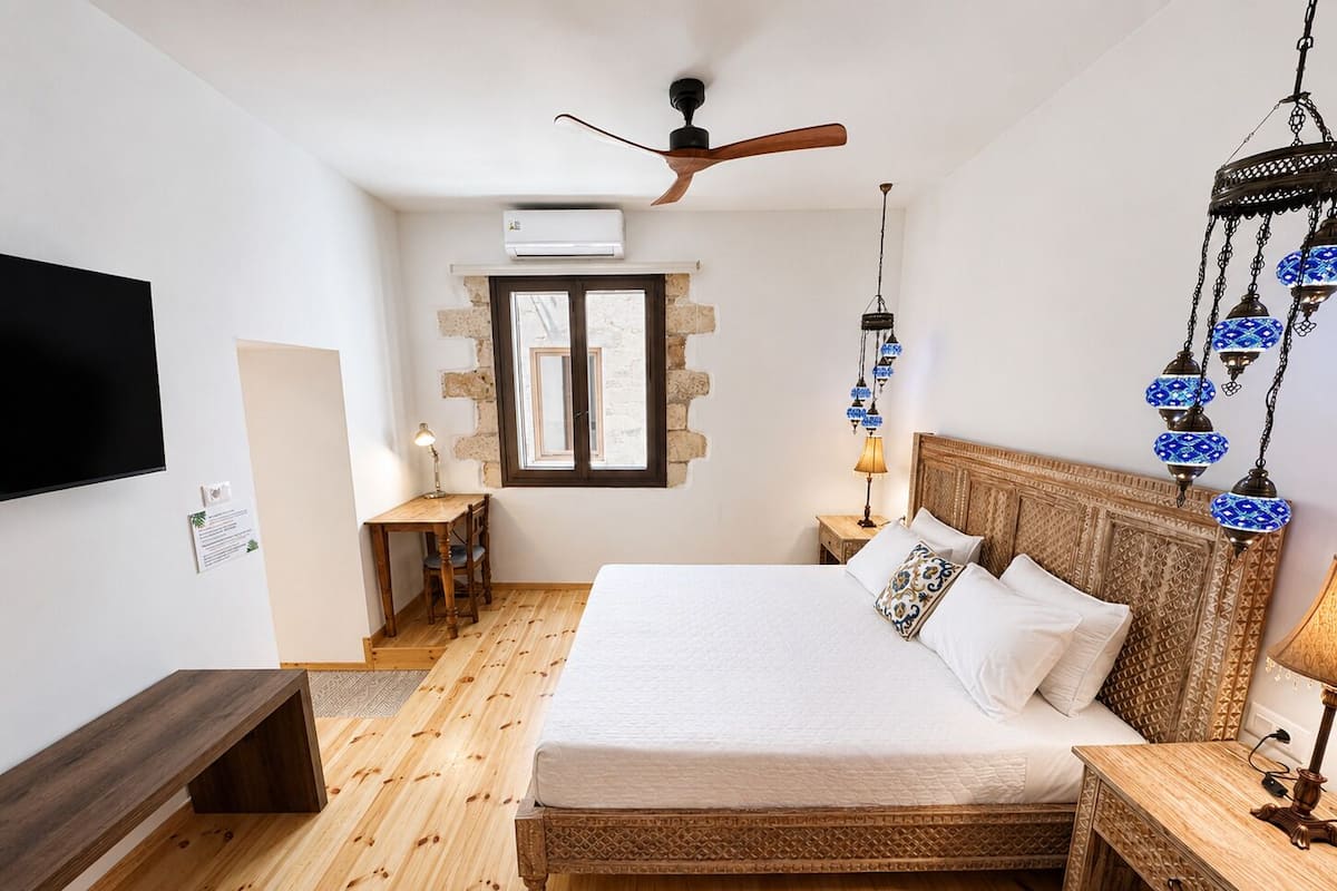 Um quarto com cama de madeira, ventoinha no teto, televisão, janela e luminária pendente.